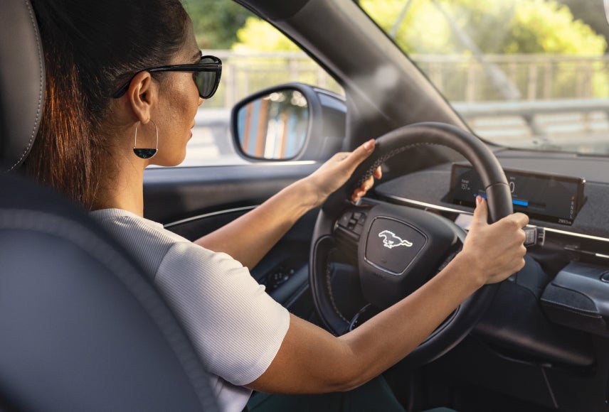 An over the shoulder view of a woman behind the wheel of a Mustang Mach-E® SUV driving over a bridge.