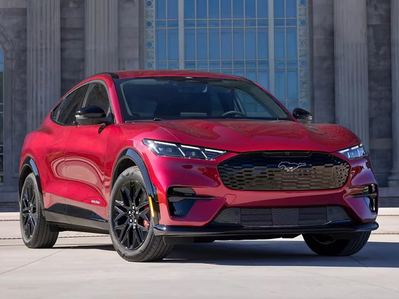 A red 2025 Ford Mustang Mach-E® SUV parked in front of an ornate concrete building with pillars and stained glass.